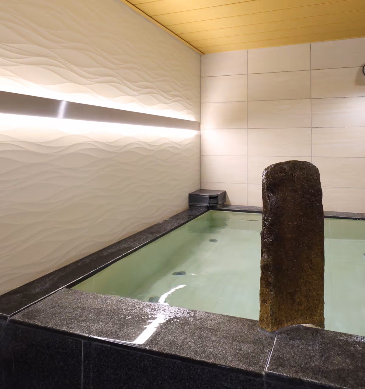 Indoor rectangular stone bath with clear water and a tall textured stone in the corner of the bath.