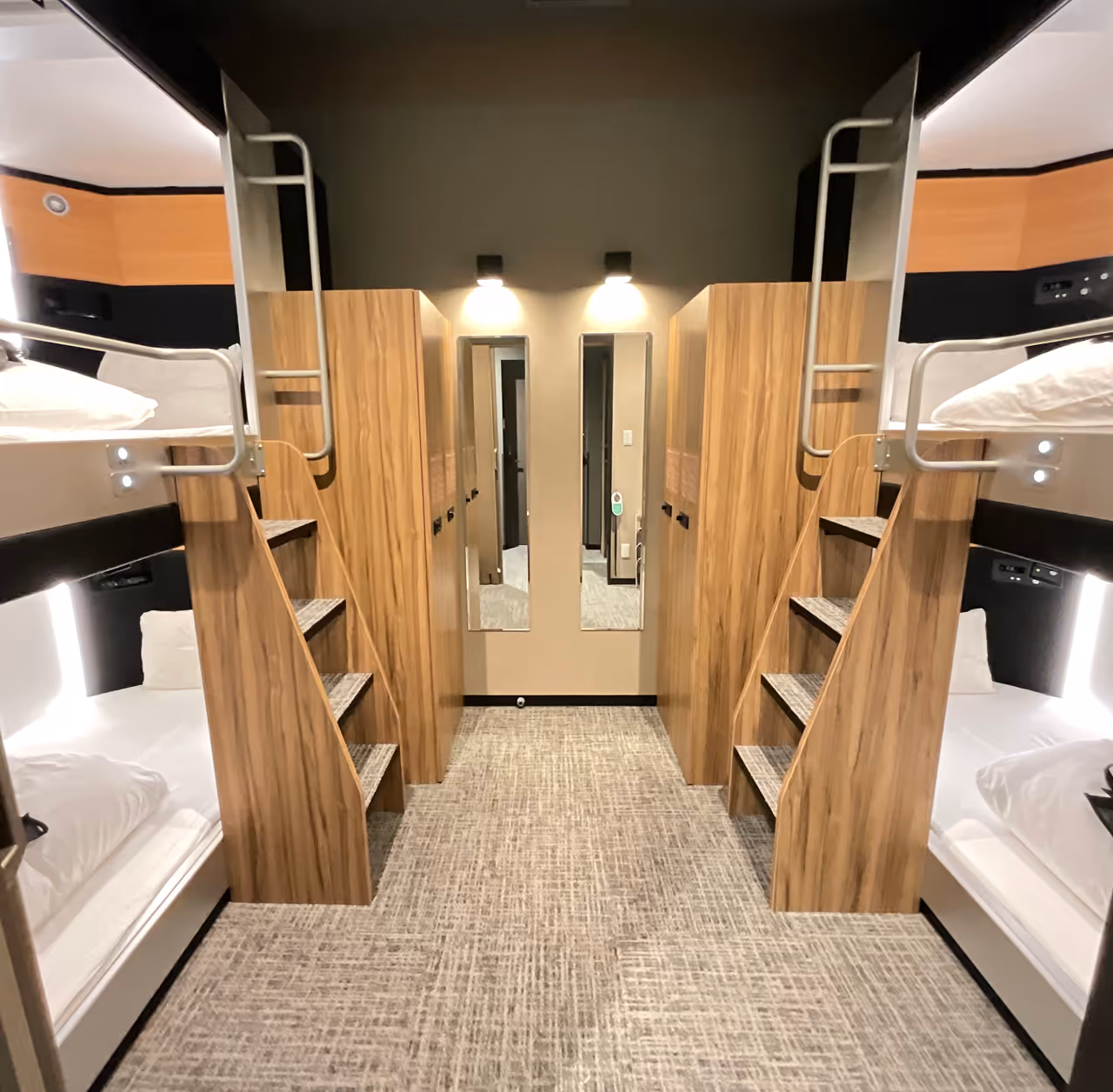 Modern dormitory room with two sets of wooden bunk beds, each with stairs and individual reading lights, and two vertical mirrors on the back wall.