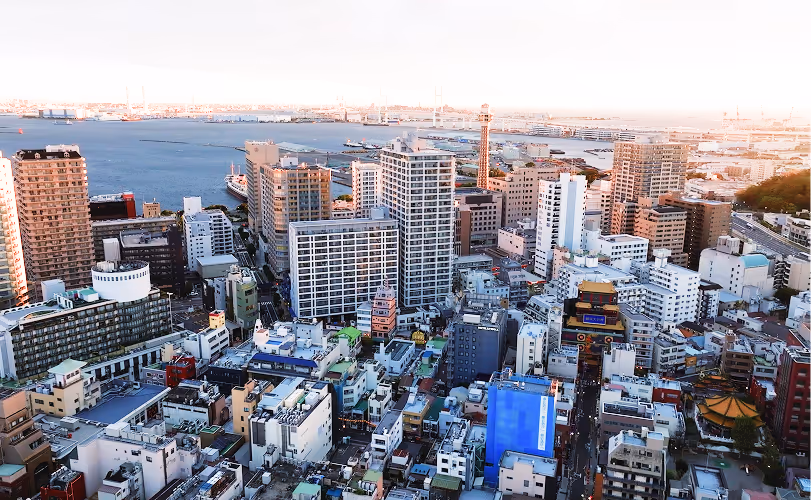 Aerial view of a cityscape with numerous high-rise buildings and a large body of water in the background under a bright sky.