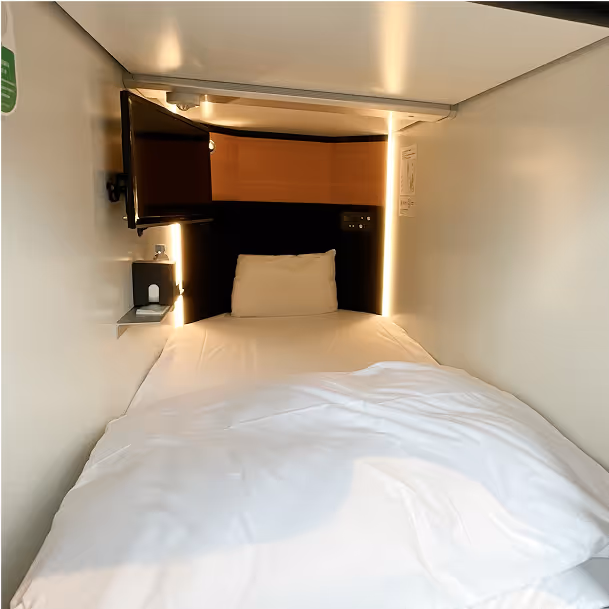 Small, modern capsule hotel room with a single bed, white bedding, a pillow, wall-mounted TV, and ambient lighting.