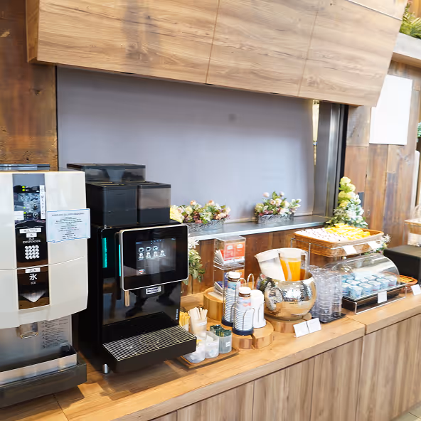 Self-service drink station with coffee machines, disposable cups, juice dispenser, and snacks on a wooden counter with floral decorations.