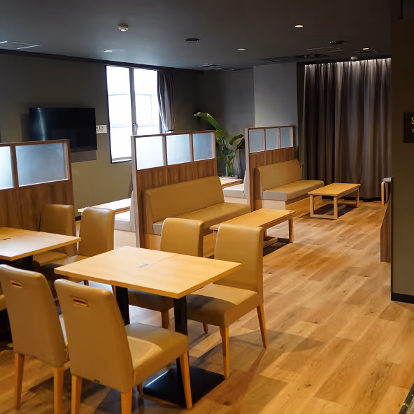 Modern dining area with beige cushioned chairs and wooden tables on hardwood flooring, featuring booth seating and a wall-mounted TV.