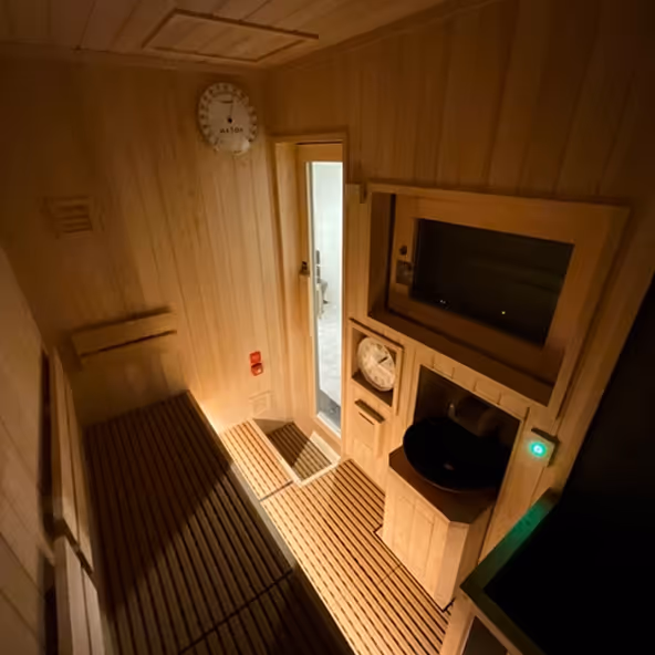 Interior of a wooden sauna with benches, a thermostat, a clock, and a door leading to a lighted room.