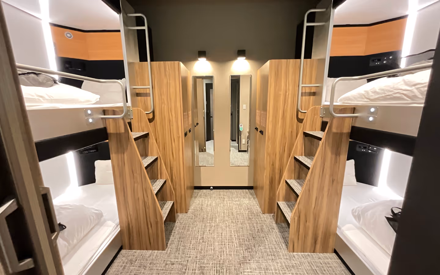 Modern dormitory room with two sets of wooden bunk beds, each with stairs and individual reading lights, and two vertical mirrors on the back wall.