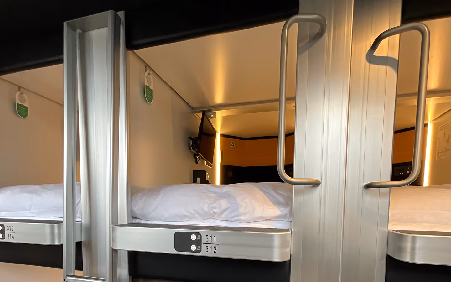 Interior of a modern capsule hotel room showing a neatly made bed with white linens, a wall-mounted TV, and ambient lighting.