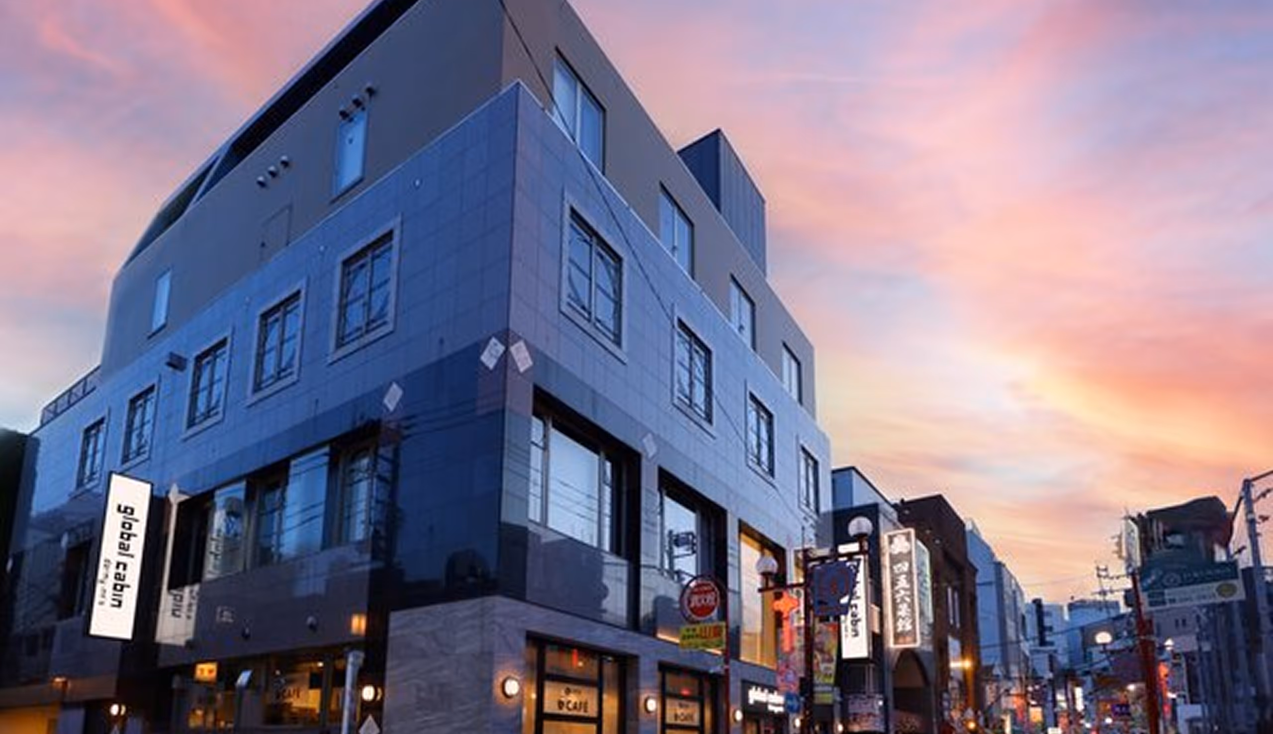 Modern multi-story building with large windows and illuminated signs at sunset on a busy urban street.