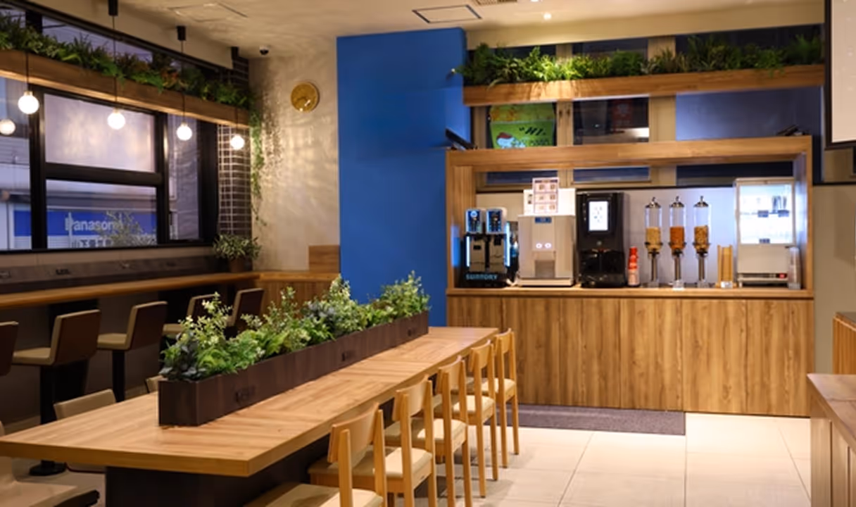 Modern lounge area with wooden tables and chairs, plants as centerpiece, beverage dispensers, and hanging lights near large windows.