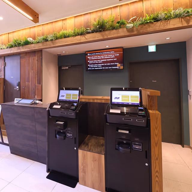 Modern hotel reception area with two black self check-in kiosks and a wooden counter adorned with green plants above.