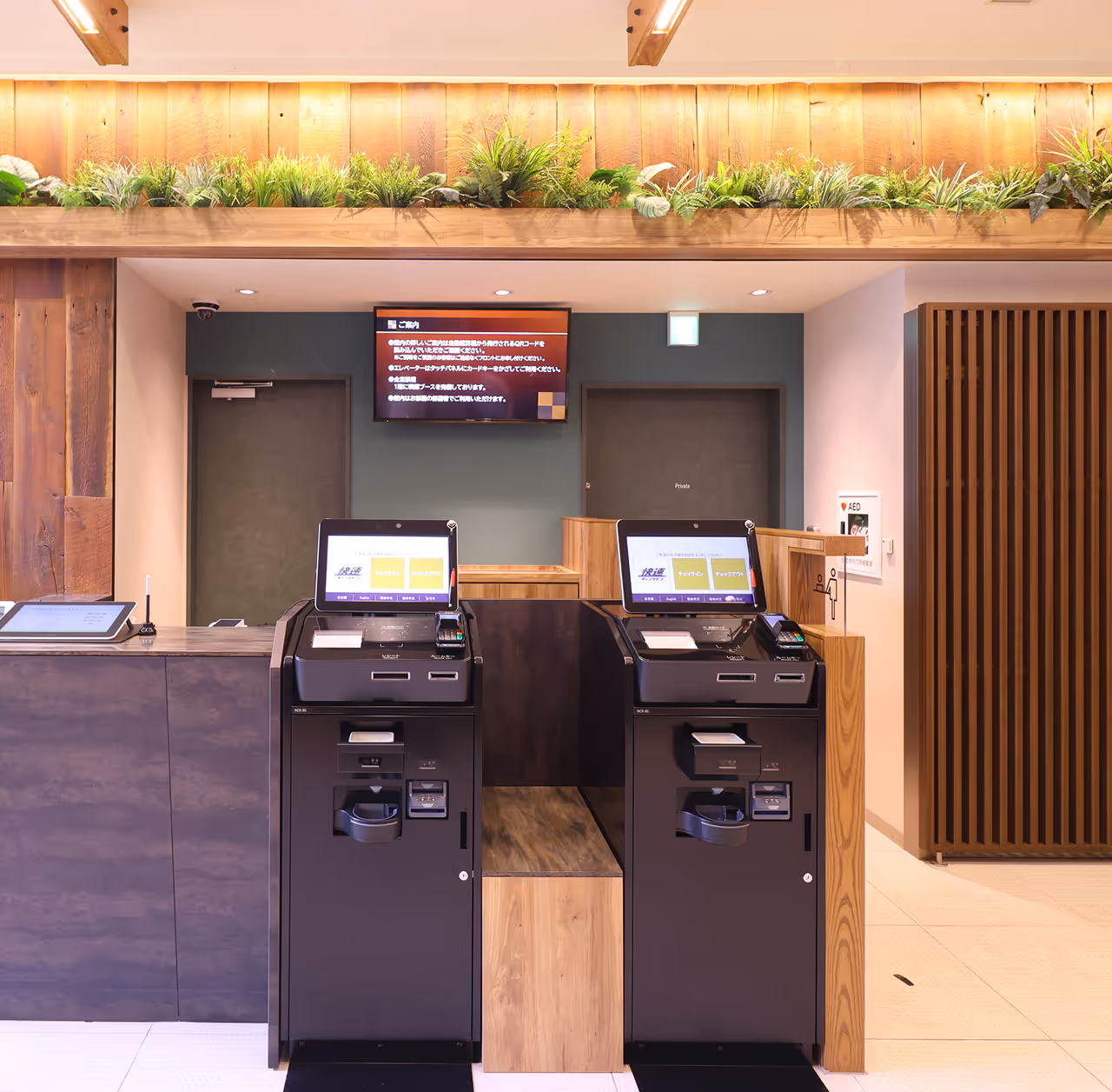 Modern self-service check-in kiosks with touchscreen monitors in a clean, well-lit reception area featuring wood accents and green plants.