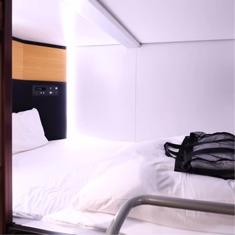 Small modern bed with white bedding and a black backpack resting on it in a compact, light-colored room.