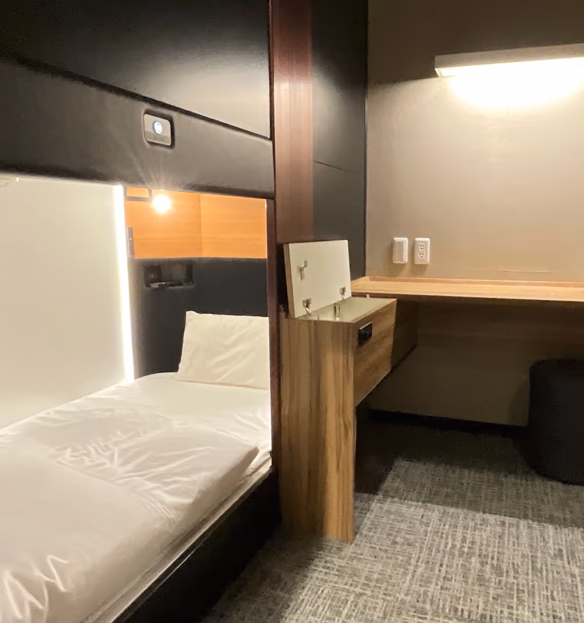 Compact room with a bed on the left and a wooden desk with an open storage compartment on the right under a wall light.