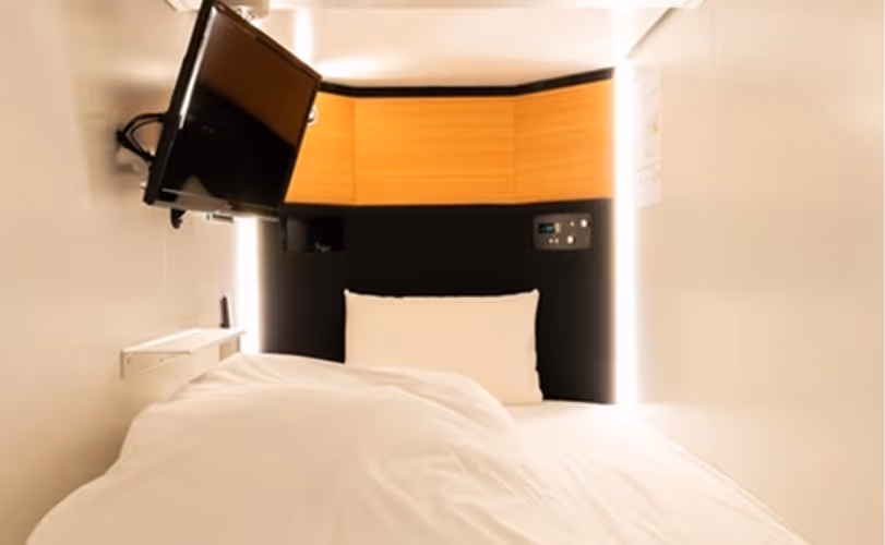 Compact, modern capsule hotel room with white bedding, wall-mounted TV, and illuminated wooden panel.