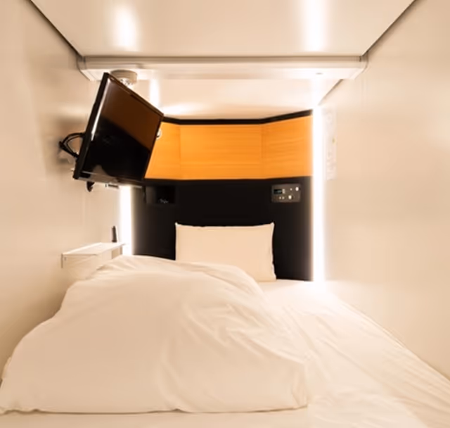 Compact capsule hotel room with a bed, white bedding, a mounted flat-screen TV, and illuminated wooden panel at the headboard.