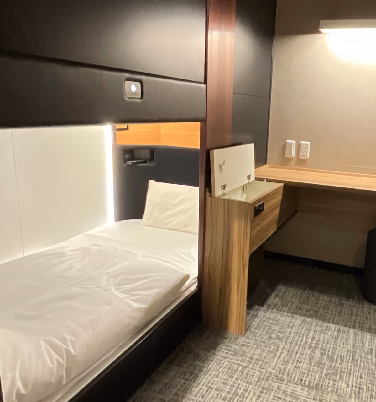 Small modern room with a single bed, white bedding, a wooden nightstand with an open panel, and a wooden desk under a wall light.
