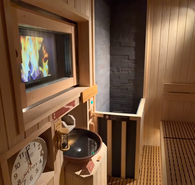 Interior of a wooden sauna with a clock, hourglass, and a bowl, and a visible fireplace through a glass window.