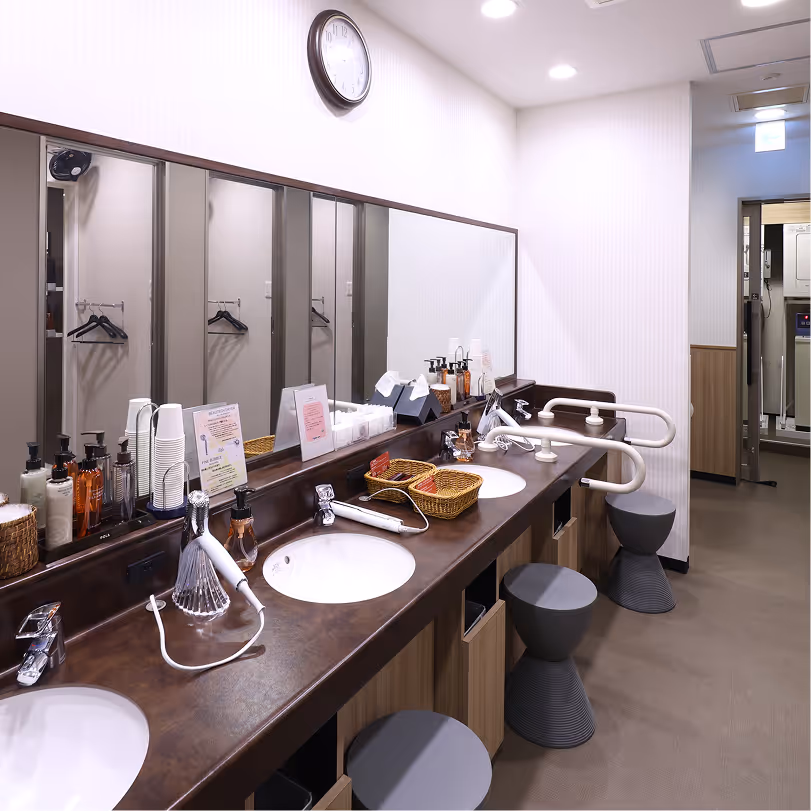 Clean public restroom with three sinks, hairdryers, liquid soap dispensers, round stools, and a large mirror under a wall clock.