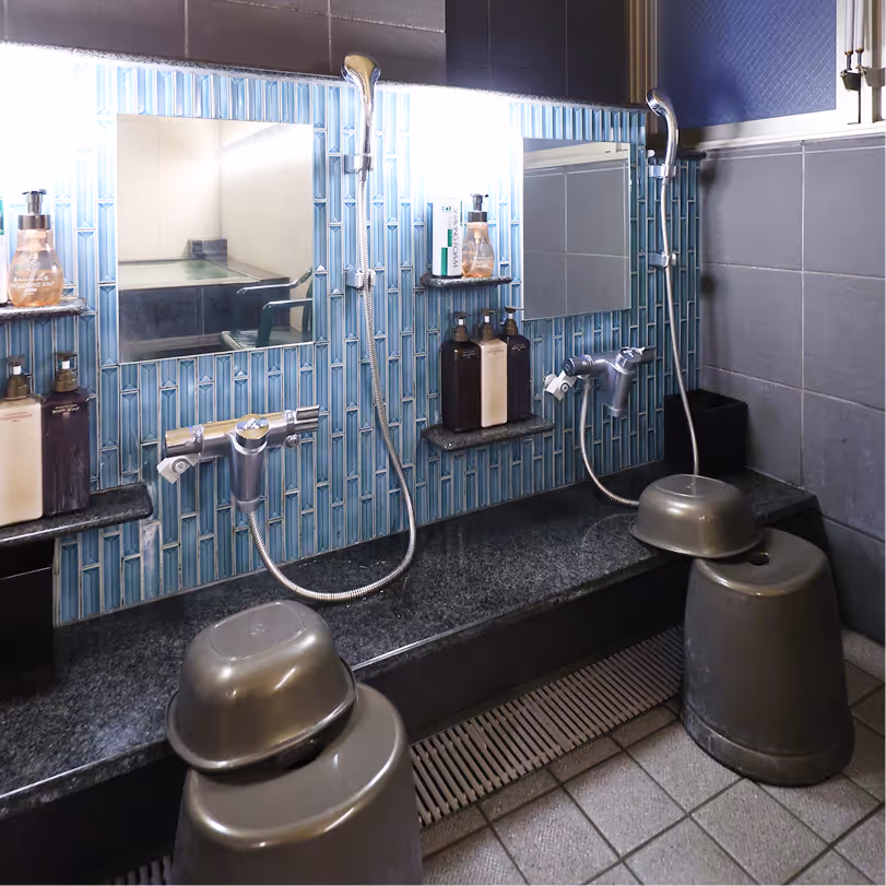 Traditional Japanese-style washing area with stools, handheld showerheads, mirrors, and toiletries on a tiled wall.